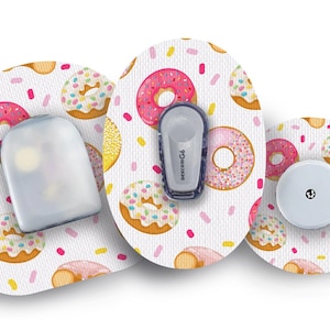 Może przedstawiać: Trzy okrągłe, białe plastry medyczne z wzorem pączków. Plastry mają różowe, żółte i białe pączki z posypką. Jeden plaster ma przymocowany do niego mały, szary, medyczny przyrząd. Urządzenie ma napis "dexcom G6".