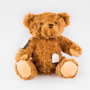May include: A brown teddy bear with white patches on its arms and chest. The bear is sitting on a white surface.