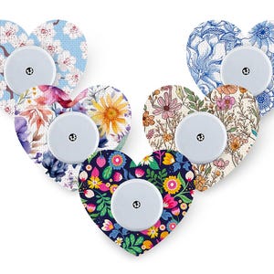 Puede incluir: Cinco parches adhesivos en forma de corazón para sensores médicos, cada uno con un patrón distinto: flores de cerezo azul claro, floral azul y blanco, flores silvestres multicolores, estampado de pradera y fondo azul oscuro con flores vivas.