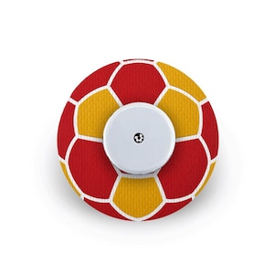 May include: A circular adhesive patch featuring a red and yellow hexagonal pattern resembling a football. The centre of the patch has a smooth, white, circular plastic device with a small black icon printed in the middle of the surface.