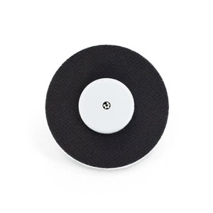 Puede incluir: Un electrodo adhesivo redondo negro con un círculo central blanco y un pequeño punto negro en el centro.