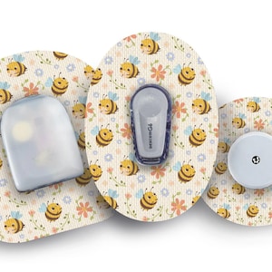 以下が含まれることがあります： 漫画の蜂と花の模様がついた丸い粘着パッチが3つ。パッチはベージュで、黄色い蜂とピンク、青、オレンジの花が描かれている。中央のパッチには、小さな丸い白いセンサーがあり、中央に黒い点がある。左のパッチには、透明な長方形のデバイスがあり、「dexcom G6」という文字が印刷されている。