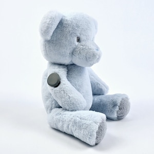 May include: A light blue plush teddy bear with a grey button on its arm. The bear is sitting with its legs crossed and its head tilted to the side.
