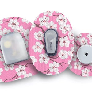 May include: Three medical device components with a pink floral design. The components include a sensor, a transmitter labeled "Dexcom G6", and a round white device. The floral pattern features white cherry blossoms on a pink background.