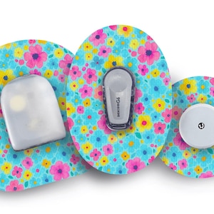 Puede incluir: Tres dispositivos médicos con parches adhesivos con estampado floral. Los parches son de color azul claro con flores rosas, amarillas y moradas. Los dispositivos incluyen un sensor blanco, un sensor gris y un dispositivo redondo blanco.