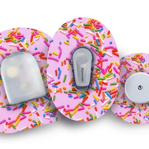 Könnte beinhalten: Drei runde medizinische Geräte-Pflaster mit rosa Hintergrund und bunten Streuseln. Die Pflaster sind zum Tragen auf der Haut konzipiert. Das mittlere Pflaster hat einen kleinen weißen Kreis mit einem schwarzen Punkt in der Mitte. Das linke Pflaster hat eine transparente Plastikabdeckung. Das rechte Pflaster hat ein kleines graues Gerät mit dem aufgedruckten Text "dexcom G4".