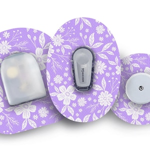 Pode incluir: Três adesivos com padrão floral branco e roxo para um dispositivo médico. Os adesivos são redondos e têm um design floral branco sobre um fundo roxo. Um adesivo tem um pequeno sensor redondo cinza, outro tem um pequeno sensor retangular cinza e o terceiro tem um sensor retangular cinza maior com a palavra "FreeStyle" impressa nele.