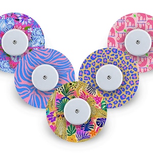 Op de afbeelding: Vijf ronde zelfklevende pleisters voor medische sensoren, elk met een uniek patroon: blauw-witte tijgers met roze bloemen, blauw-roze zebrastrepen, geel-blauwe luipaardvlekken, roze luiaards en een kleurrijke luipaard met tropische bladeren.