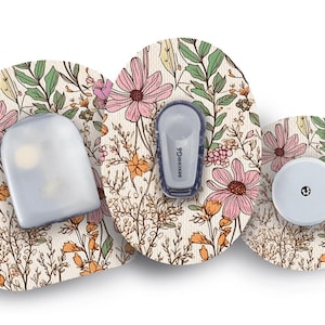 Op de afbeelding: Een set van drie medische apparaten, waaronder een sensor en een zender, met een bloemenontwerp. De apparaten zijn versierd met roze bloemen, groene bladeren en oranje accenten op een crèmekleurige achtergrond. De apparaten zijn voor medisch gebruik.