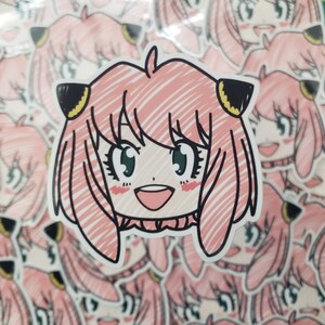 May include: A pink and white sticker with a cartoon drawing of a girl with pink hair, green eyes, and a big smile. The girl is wearing a black collar and has yellow cat ears.