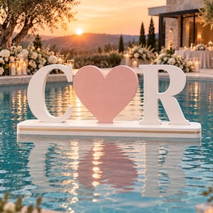 May include: A white letter C and a white letter R flanking a central pink heart shape, all mounted on a rectangular white base with a thin gold horizontal stripe, floating on the surface of a swimming pool at sunset.
