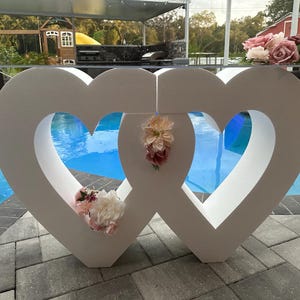 Pode incluir: Duas grandes estruturas em forma de coração de espuma branca com centros recortados, colocadas lado a lado. Os corações são decorados com flores rosa e brancas. Os corações estão em frente a uma piscina.