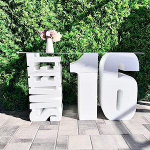 Puede incluir: Número 16 blanco de espuma y la palabra "Sweet" apilados uno encima del otro sobre una mesa de vidrio frente a una pared verde.