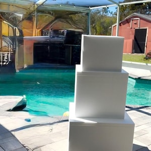 Puede incluir: Tres niveles de pastel de espuma blanca apilados uno encima del otro. Los niveles son cuadrados y están sentados en un patio junto a una piscina.