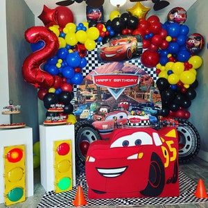 May include: A Cars-themed birthday party decoration with red, yellow, and blue balloons, a checkered backdrop, and a large cutout of Lightning McQueen. The backdrop features the text "Happy Birthday".