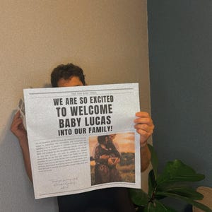 May include: A person holding a newspaper announcement that reads "We are so excited to welcome Baby Lucas into our family!" with a photo of a couple holding a baby in the bottom right corner.