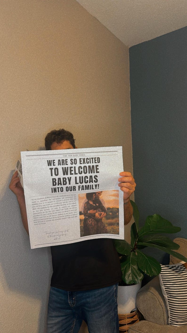 May include: A person holding a newspaper announcement that reads "We are so excited to welcome Baby Lucas into our family!" with a photo of a couple holding a baby in the bottom right corner.