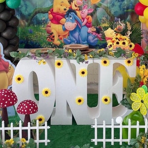 Puede incluir: Letras de espuma blanca que deletrean "ONE" decoradas con girasoles amarillos. Las letras están sobre una mesa con un telón de fondo temático de Winnie the Pooh. También hay globos amarillos y rojos, plantas verdes y vallas blancas.