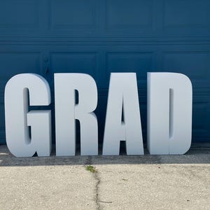 May include: Large, white, freestanding letters spelling out the word "GRAD" are displayed on a concrete surface. The letters are positioned in front of a blue garage door, creating a celebratory theme.