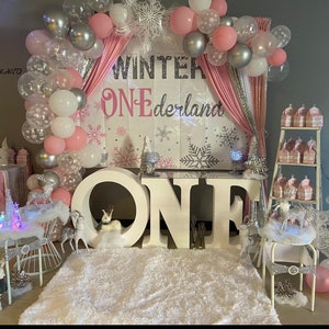 Könnte beinhalten: Eine Winterwunderland-Motto-Geburtstagsparty zum ersten Geburtstag mit weißen, rosa und silbernen Ballons, einer Kulisse mit den Worten "Winter One-derland" und großen weißen Buchstaben, die "ONE" buchstabieren.