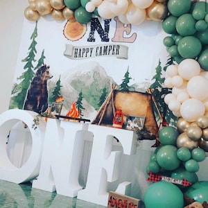 Puede incluir: Letras de espuma blanca que deletrean "ONE" con un telón de fondo de temática boscosa que presenta un oso, una tienda de campaña y una fogata. El telón de fondo tiene el texto "Happy Camper". Hay un arco de globos verde, dorado y crema en el fondo.