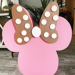 Puede incluir: Un recorte de cartón rosa y dorado de la cabeza de Minnie Mouse con un gran lazo. El lazo es marrón con lunares blancos.