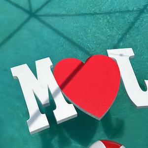 Puede incluir: Una piscina con un corazón rojo, letras blancas que deletrean "MOM" y una pelota de playa amarilla, blanca y azul flotando en el agua.