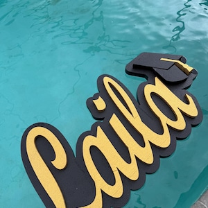 Könnte beinhalten: Ein Graduation-Schwimmring mit dem Namen "Laila" in goldenen Buchstaben mit schwarzen Umrissen. Der Schwimmring befindet sich auf einer blauen Pool-Oberfläche.