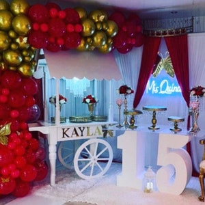 Puede incluir: Un carro de postres blanco con la palabra "KAYLA" escrita en el lateral. El carro está decorado con globos rojos y dorados y tiene un gran "15" blanco delante. El carro está preparado para una fiesta con un cartel que dice "Mis Quince" en luces de neón.
