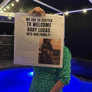 May include: A woman in a green polka dot dress holds a newspaper announcing the birth of "Baby Lucas" with the headline "We are so excited to welcome Baby Lucas into our family!" The newspaper features a photo of a couple holding hands with the woman's hand on her stomach.