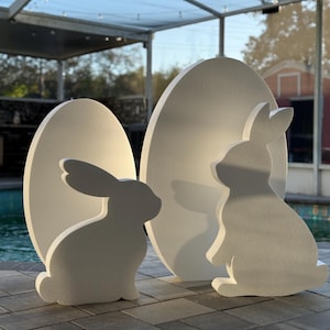 May include: Three white Easter decorations: two bunny silhouettes and an egg-shaped backdrop. The bunnies are in different poses, one sitting and one standing. The decorations are placed on a stone patio.