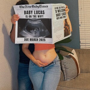 May include: A couple holds up a newspaper announcing the pregnancy of "Baby Lucas" with a due date of March 2025. The newspaper features an ultrasound image and the headline "Baby Lucas is on the way!"