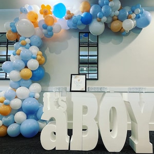 Puede incluir: Letras de espuma blanca que deletrean "It's a Boy" sobre una mesa de vidrio con un arco de globos en el fondo. El arco de globos está hecho de globos azules, blancos y naranjas.