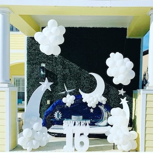 Puede incluir: Decoraciones de globos blancos en forma de nubes y estrellas, con un gran número 16 blanco y la palabra "SWEET" en letras blancas. Las decoraciones están instaladas frente a una pared verde con un fondo azul.