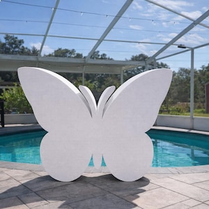 May include: A large, white butterfly sculpture stands near a swimming pool. The butterfly has detailed wing shapes and is positioned on a stone patio. The pool's water is a vibrant blue, and the background includes trees and a red shed.