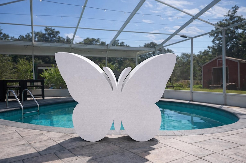 May include: A large, white butterfly sculpture stands near a swimming pool. The butterfly has detailed wing shapes and is positioned on a stone patio. The pool's water is a vibrant blue, and the background includes trees and a red shed.