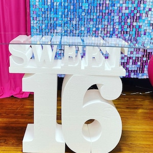 Puede incluir: Letras blancas de espuma que deletrean "Sweet Sixteen" sobre una mesa de vidrio con un fondo azul y morado brillante.