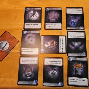 May include: A tabletop card game with illustrated cards featuring robots and abstract designs. The cards have text descriptions and numbers. A brown card with a metallic design is also present.
