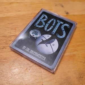 May include: A deck of cards in a clear plastic case. The card design features the word "BOTS" in blue, with a robot illustration and the text "BY ZAC BAUERMEISTER". The background is dark gray.
