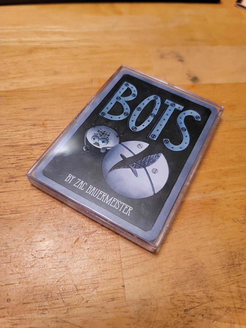 May include: A deck of cards in a clear plastic case. The card design features the word "BOTS" in blue, with a robot illustration and the text "BY ZAC BAUERMEISTER". The background is dark gray.