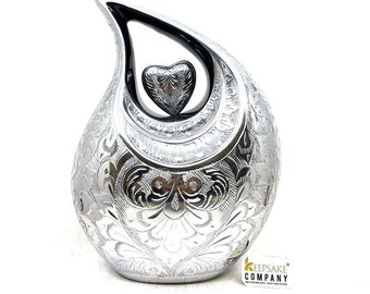 May include: Silver teardrop-shaped cremation urn with an engraved floral design. The urn has a small heart-shaped opening on the top with a silver heart inside.