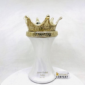 May include: White ceramic urn with a gold crown on top. The urn is engraved with the name "Jasmine Elizabeth Rodkey" and the dates "2022 - 2023". The urn is made by the Keepsake Company.