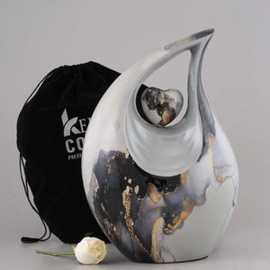 May include: A teardrop-shaped urn with a heart-shaped insert. The urn features a white, gray, black, and gold marbled design. A black drawstring bag and a white rose are also in the image.