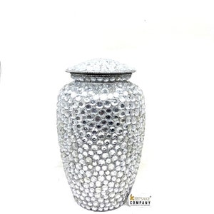 May include: A silver cremation urn with a lid covered in clear rhinestones. The urn is about 12 inches tall and has a round shape.