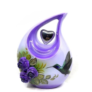 May include: A teardrop-shaped urn in shades of lavender and white, featuring a silver heart and a hummingbird design. The urn is adorned with purple roses and green leaves, creating a serene and elegant aesthetic.
