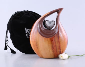 Puede incluir: Una urna funeraria de madera en forma de lágrima con un acabado de grano de madera marrón claro pulido y un recorte interior marrón oscuro. Un pequeño emblema plateado en forma de corazón está centrado en la abertura. Una bolsa negra y una rosa blanca.