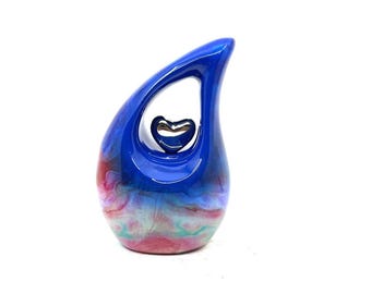 May include: A blue teardrop-shaped decorative object with a glossy finish. The object has a heart-shaped cutout and a smaller heart-shaped accent inside. The base features a blend of pink, blue, and green hues.