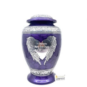 May include: A deep purple cremation urn with a silver lid and base featuring intricate floral engravings. The center displays a silver heart-shaped plaque with angel wings, engraved with the text In Loving Memory Mary Ann Creasy 08 08 1948 12 27 2022.