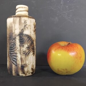 May include: A tall, cylindrical ceramic bottle with a textured, wood-like appearance. The bottle features a circular, carved design and a light-coloured stopper. A yellow and red apple sits next to the bottle.