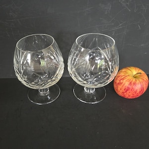May include: Two clear crystal brandy snifters with diamond-cut patterns. The glasses sit on a black surface next to a red and yellow apple. The snifters have a rounded bowl and a short stem.